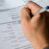 Dłoń pisze na formularzu "Zgłoszenie pobytu stałego", wpisując nazwisko. Dokument ten jest kluczowy do uzyskania tytułu prawnego do lokalu.
