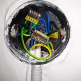 Widok wnętrza puszki elektrycznej z przewodami i złączkami. Trwa mostkowanie gniazdka, łącząc przewody niebieski, zielono-żółty i brązowy.