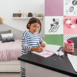 Dziewczynka rysuje w swoim małym pokoju dla dziecka. Na biurku leżą kolorowe kamyczki i papier.