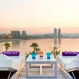 Dos tumbonas blancas con toallas moradas de W Hotels, una mesa con vasos y velas, y vistas al mar y la ciudad de Barcelona al atardecer.