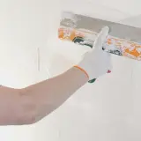 Mano con guanto bianco stende stucco murale su una parete bianca con una spatola arancione.