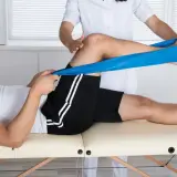 Fizjoterapeuta pomaga pacjentowi w ćwiczeniach mięśnia czworogłowego uda z użyciem taśmy oporowej.