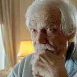 En äldre svensk skådespelare med vitt hår och mustasch funderar djupt, med handen mot hakan.