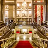 Elegancki foyer z marmurowymi schodami, złotymi balustradami i kryształowym żyrandolem. Czerwony dywan prowadzi do przestronnego holu.