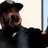 Raper Ice Cube w czapce z daszkiem "ICE CUBE" śpiewa do mikrofonu, gestykulując ręką. Jego styl to klasyczny diss.