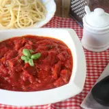Un tazón blanco lleno de salsa marinara casera, decorada con albahaca fresca, junto a espaguetis cocidos.