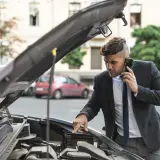 Mężczyzna w garniturze dzwoni po pomoc drogową, wskazując na otwartą maskę samochodu.