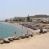 Słoneczna plaża na wyspie Kos z białymi hotelami, parasolami i turystami kąpiącymi się w morzu.
