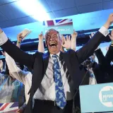 Jubelnde Menge bei einer Wahl in England. Ein Mann mit erhobenen Armen und einem breiten Lachen im Vordergrund, umgeben von Union-Jack-Flaggen und "Reform UK"-Schildern.