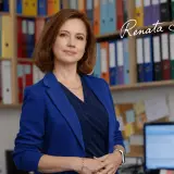 Kobieta w niebieskiej marynarce stoi przed regałami z kolorowymi segregatorami.