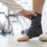 Dłoń zakłada ściągacz na kostkę, by zapewnić jej stabilizację. W tle widać rehabilitacyjny sprzęt.