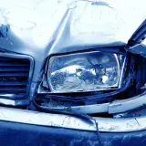 Il cofano e il faro di un'auto sono gravemente danneggiati, con lamiera accartocciata e vetro rotto. Sembra il risultato di **atti vandalici**.