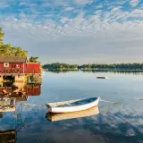 En vit båt ligger förtöjd vid en röd sjöbod. Kanske är det en båt till Drottningholm, redo för en lugn tur på vattnet.