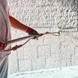 Pracownik w rękawicach nakłada obrzutkę cementową na ścianę za pomocą agregatu.