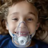 Uśmiechnięte dziecko z kręconymi włosami w masce do inhalacji.