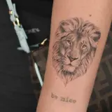 Tatuaż znak zodiaku lew na ramieniu, z napisem "be nice" poniżej.
