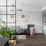 Nowoczesny salon z zieloną ścianą z kwiatów, szarą sofą, fotelem i przeszkloną witryną.