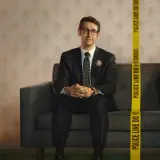 Dwóch mężczyzn siedzi na kanapie. Jeden, ubrany w garnitur, to **the good cop**. Obok niego, za taśmą policyjną, drugi mężczyzna uśmiecha się.