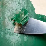 Metalowa szpachelka zdziera starą, zieloną farbę, odsłaniając podkład.