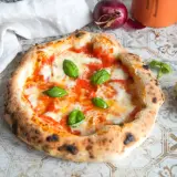 Eine authentische Pizza Napoletana mit frischem Basilikum, geschmolzenem Mozzarella und Tomatensauce. Daneben ein Stück Parmesan und eine rote Zwiebel.