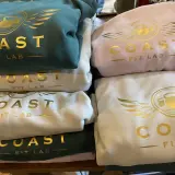 Stosik bluz klubowych w kolorach zielonym, różowym i kremowym, z złotym logo "COAST FIT LAB".