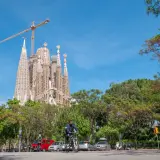 Majestatyczna La Sagrada Familia w Barcelonie, wciąż w budowie, góruje nad zielonym parkiem i ulicą, gdzie rowerzysta przemierza drogę.