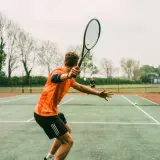 Młody tenisista w pomarańczowej koszulce i czarnych spodenkach wykonuje potężny serwis, celując w klasyczny wielki szlem.