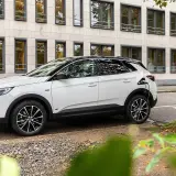 Biała hybryda plug-in ładuje się przy stacji ładowania. Nowoczesny samochód stoi na tle budynku z wieloma oknami.