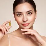 Mujer sonriente sostiene una cápsula dorada de vitamina e, símbolo de cuidado y bienestar para una piel radiante.
