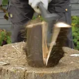 Ein Mann spaltet mit einem Keil einen Baumstamm. Holzsplitter fliegen durch die Luft.