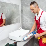 Fachowiec w czerwonym ogrodniczkach przygotowuje się do usuwania starego silikonu z łazienki, trzymając pistolet do silikonu.