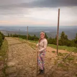 Uśmiechnięta turystka z kijkami na kamienistej ścieżce, podziwia widok na góry. Widać, ile się wchodzi na Śnieżkę.