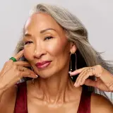 Mujer con cabello plateado, luciendo un tinte o mechas naturales, con labios rojos y joyas doradas.