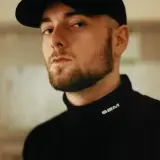 Chudy Bedoes w czarnej bluzie z golfem i czapce z daszkiem, z charakterystyczną brodą.