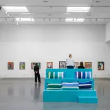 I en konsthall sitter besökare på en turkos scen med kuddar, omgivna av färgglada tavlor. Kanske är de där för att inspireras, för att vara en musa för konstnären.