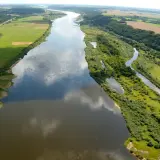 Szeroka rzeka Nemen meandruje przez zielone pola i lasy, odbijając chmury na swojej tafli.