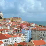 One day in Lisbon, a panorama of colorful buildings with red-tiled roofs stretches towards the shimmering blue sea under a cloudy sky.