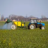 Niebieski traktor z żółtym zbiornikiem wykonuje oprysk na polu zielonych roślin. W tle widać przemysłowe budynki.