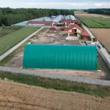 Zielona hala namiotowa na terenie gospodarstwa. Obok budynki mieszkalne i gospodarcze, otoczone polami uprawnymi. Grunty rolne zabudowane.