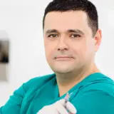 Lekarz w zielonym fartuchu trzyma strzykawkę. W tle widać elementy gabinetu. Wiele pozytywnych klinika ambroziak opinie potwierdza jego profesjonalizm.