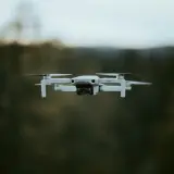 Biały dron DJI Mini 2 w locie, z widocznym obiektywem kamery. Tło to rozmyty las i niebo.