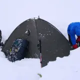 Osoba w niebieskiej kurtce i czerwonych rękawiczkach przygotowuje namiot w śniegu. Obok leżą plecaki i kijki trekkingowe, które pomagają dobrać kijki trekkingowe do wzrostu.