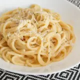 Pyszne spaghetti carbonare, posypane startym parmezanem i świeżo mielonym pieprzem, w białej misce na wzorzystym obrusie.