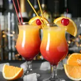 Dwa drinki z wódką w kieliszkach z pomarańczą i wiśnią, na tle baru.