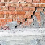 Uszkodzona ceglana ściana z widocznym pęknięciem w betonie, sugerującym podcinanie fundamentów.