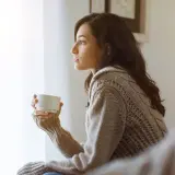 Kobieta w swetrze z kubkiem w dłoni, zamyślona, jakby czekała na coś ważnego, co symbolizuje moment, kiedy znajomość przeradza się w związek.