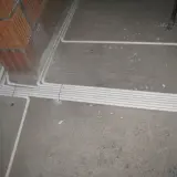 Instalacja elektryczna w podłodze, przygotowana zgodnie z przepisami. Białe rury układają się wzdłuż betonowej posadzki, obok ceglanej ściany.