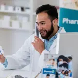 Uśmiechnięty farmaceuta rozmawia przez telefon, trzymając pudełko leku. Zastanawia się, ile zarabia farmaceuta, pracując w aptece.