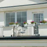 Białe okna na jasnej, teksturowanej elewacji budynku. Balkon z czarną balustradą i donicami z kwiatami.