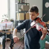 Chłopiec gra na skrzypcach z pasją, dźwięki rozbrzmiewają **fortissimo**. W tle inni uczniowie ćwiczą muzykę.